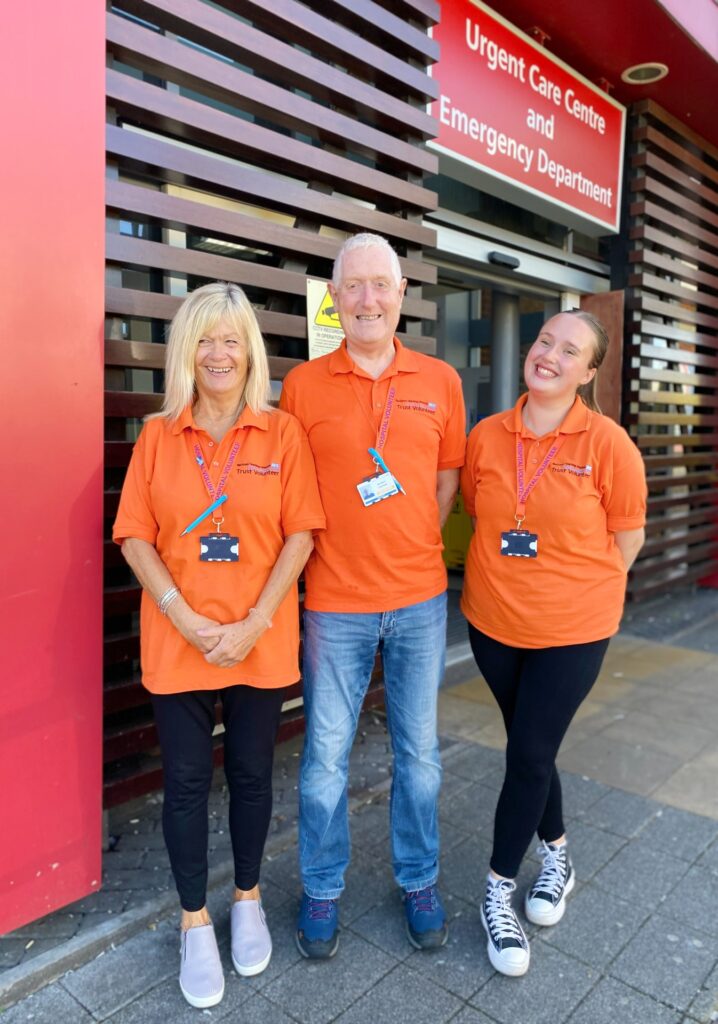 Three people in orange polo shirts standing at the Emergency Department entanace